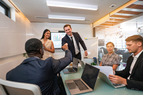 Coworkers sitting around a table smiling, and one is giving a fist bump to someone standing up